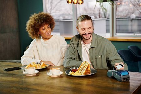 Une femme afro-américaine et un homme assis à une table, dégustant des assiettes de nourriture dans un café moderne.