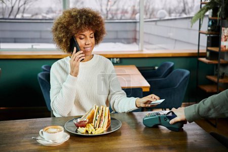 Photo for An African American woman engaged in a conversation over a cell phone while seated at a table in a modern cafe. - Royalty Free Image