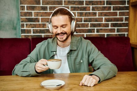 A man of African descent sits at a cafe table, sipping coffee and listening to music through headphones.