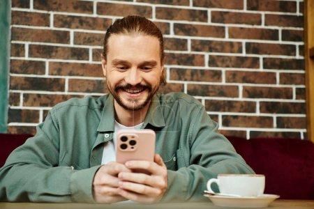 happy man is seated at a table in a modern cafe, focused on using his cellphone.