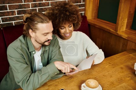 An African American woman and man sit at a table, choosing meal in menu in a modern cafe.