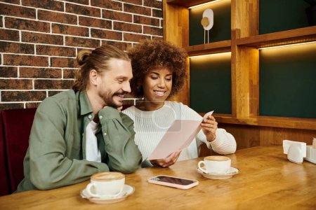 Téléchargez les photos : Une femme afro-américaine et un homme assis à une table, concentrés sur un menu dans un cadre de café moderne. - en image libre de droit