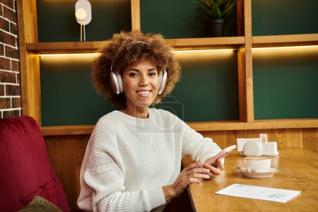 Eine afroamerikanische Frau sitzt an einem Tisch in einem modernen Café, vertieft in den Rhythmus ihrer Kopfhörer.