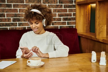Téléchargez les photos : Une Afro-Américaine s'assoit à une table dans un café moderne, profondément absorbée par son téléphone portable. - en image libre de droit