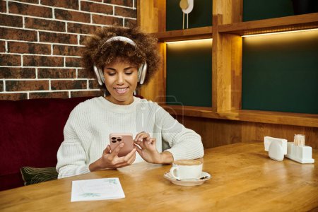 An African American woman sits at a table in a modern cafe, wearing headphones and immersing herself in her own little world.