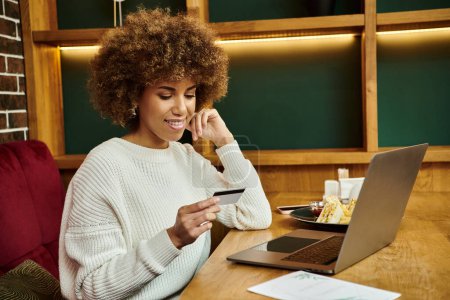 A modern African American woman sits in a cafe, focusing intently on her credit card near laptop