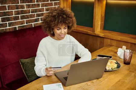 Foto de Una mujer afroamericana se sienta en una mesa en un café moderno, enfocada en la pantalla de su computadora portátil. - Imagen libre de derechos