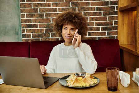 Eine afroamerikanische Frau sitzt mit Laptop und Teller an einem Tisch und konzentriert sich auf ihre Arbeit.
