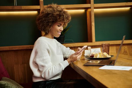 Téléchargez les photos : Une Afro-Américaine assise à une table dans un café moderne, absorbée dans son téléphone portable - en image libre de droit