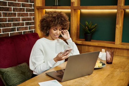 Téléchargez les photos : Une Afro-Américaine est assise à une table dans un café moderne, concentrée sur son ordinateur portable. - en image libre de droit