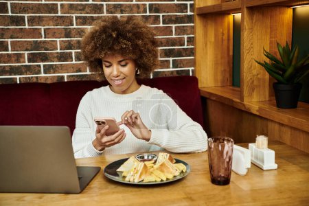 A modern African American woman sitting at a table in a cafe, engrossed in her cell phone.