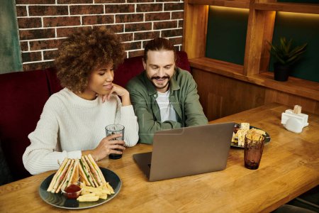 Eine afroamerikanische Frau und ein Mann sitzen an einem Tisch in einem modernen Café, beschäftigt auf dem Laptop-Bildschirm vor ihnen..
