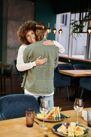 Mujer afroamericana y hombre se abrazan firmemente en la cafetería de moda, compartiendo un momento de calidez y afecto en medio de un ambiente bullicioso.