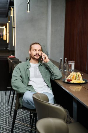 A man, seated at a table, engrossed in a phone call while surrounded by a modern cafe ambiance.