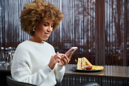 Téléchargez les photos : Une Afro-Américaine s'assoit à une table dans un café moderne, absorbée par son téléphone portable. - en image libre de droit
