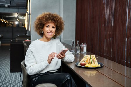A stylish African American woman sits at a table with a plate of delicious food in a modern cafe.