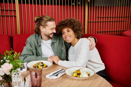 Eine afroamerikanische Frau und ein afroamerikanischer Mann genießen gemeinsam eine Mahlzeit an einem Tisch mit leckerem Essen in einem modernen Café-Ambiente.