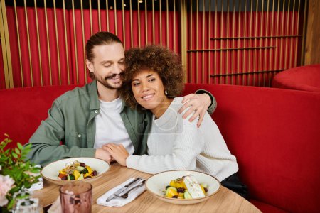 Une femme afro-américaine et un homme assis à une table, dégustant des assiettes de nourriture dans un café moderne.