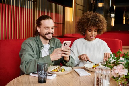 A man and woman, engrossed with a cell phone and food at a cafe table.
