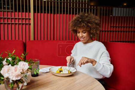 An African American woman sits at a table, enjoying a plate of food in a modern cafe.