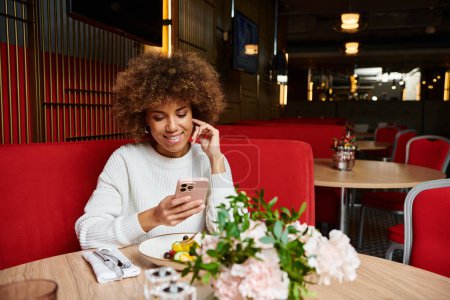 An African American woman sits at a table, engrossed in her cell phone, in a modern cafe setting.