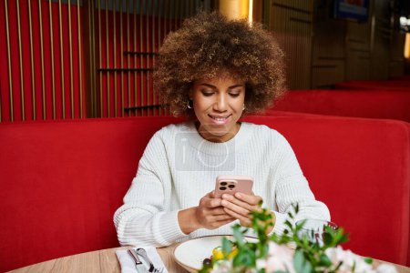 A stylish African American woman immerses in her phone while seated at a cafe table.