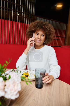 Una mujer afroamericana conversando en un celular mientras está sentada en una mesa en un café moderno.