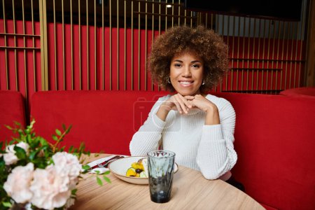 Une femme afro-américaine s'assoit à une table de café moderne, profitant gracieusement d'une assiette de nourriture délicieuse.