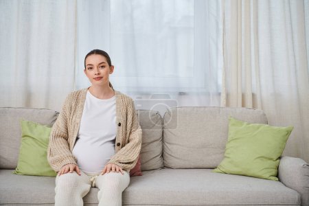 Eine schöne schwangere Frau sitzt auf einer Couch, ihre Silhouette wird vom weichen Licht eines Fensters beleuchtet.