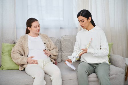 A pregnant woman and her trainer sit on a couch, attentively looking at anatomic model, parents courses.