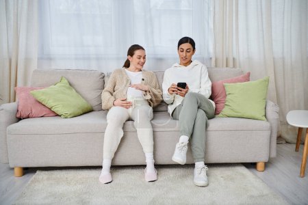 Téléchargez les photos : Un homme et une femme enceinte assis sur un canapé, absorbé dans un écran de téléphone portable, partageant probablement un moment de connexion. - en image libre de droit