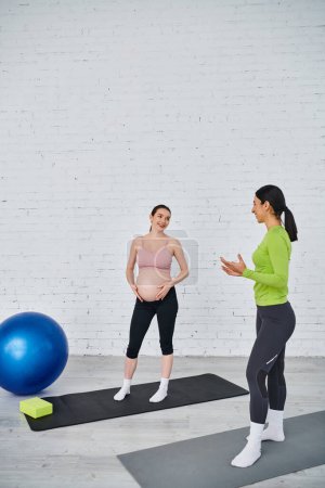 Eine schwangere Frau praktiziert Yoga auf einer Matte mit einem blauen Ball, angeleitet von ihrem Trainer während des pränatalen Kurses.