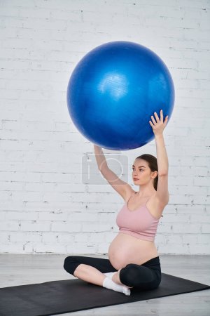 Pregnant woman in yoga pose on mat, holding blue ball during prenatal exercise session.