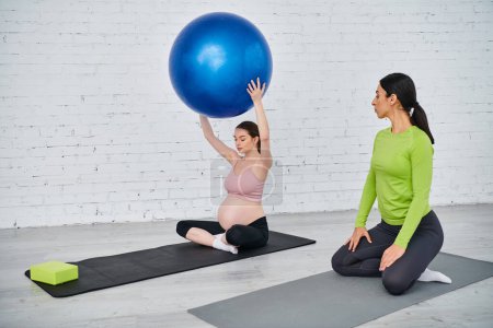 A pregnant woman sits on a yoga mat, holding a large blue ball above her head during a prenatal exercise class.