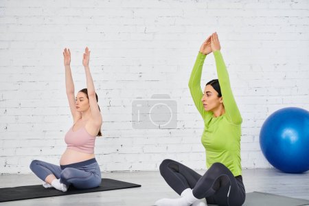 Foto de Dos mujeres practicando yoga frente a una pared de ladrillo; una está embarazada, guiada por su entrenador durante un curso de padres. - Imagen libre de derechos