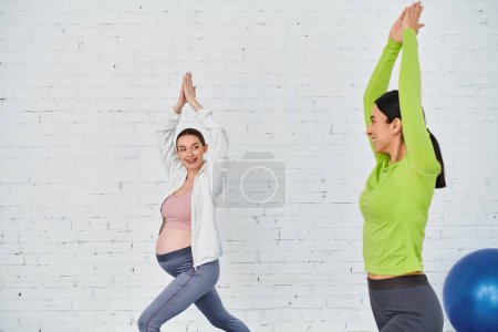 A pregnant woman exercises with her coach during a parents course, supported by another woman standing beside her.
