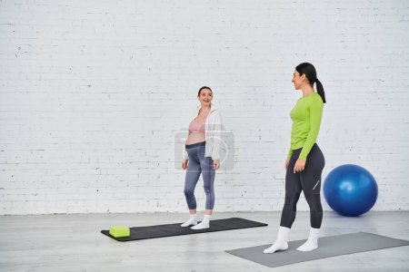 Zwei Frauen, eine schwangere, stehen auf Yogamatten in einem Fitnessstudio und erleben während eines Elternkurses einen ruhigen Moment der Achtsamkeit und Bewegung.