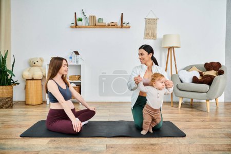 Foto de Una madre joven se sienta en una esterilla de yoga, enlazándose pacíficamente con su bebé mientras recibe orientación de un entrenador durante un curso para padres en casa. - Imagen libre de derechos