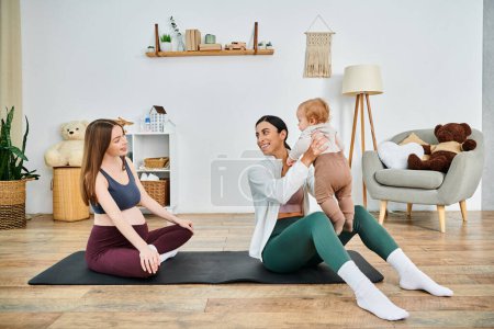 Eine junge schöne Mutter sitzt auf einer Yogamatte und hält ihr Baby auf eine ruhige und liebevolle Weise, angeleitet von ihrem Trainer bei Elternkursen.