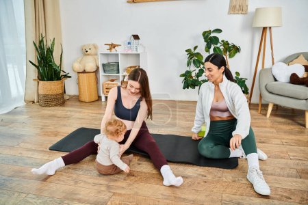 Zwei Frauen, eine gelassene junge Mutter und ihr Trainer, führen ein Baby auf einer Yogamatte in einem friedlichen häuslichen Umfeld.