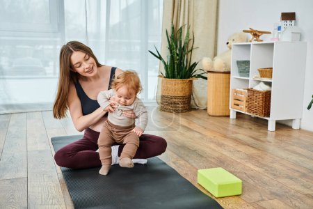 Una madre joven, sentada en una esterilla de yoga, acunando a su bebé con la guía de un entrenador en un curso para padres.