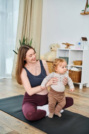 Una madre joven y hermosa se sienta en una esterilla de yoga, acunando pacíficamente a su bebé con la guía de su entrenador en los cursos de los padres.
