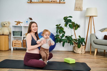 Téléchargez les photos : Une jeune mère trouve la paix sur son tapis de yoga tout en berçant son bébé avec les conseils de son entraîneur lors de cours de parents. - en image libre de droit