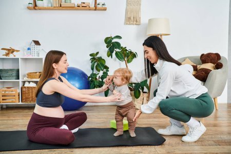 Una joven madre hermosa que se dedica a movimientos de yoga juguetones con su bebé en una esterilla, guiada por un entrenador en los cursos de padres.