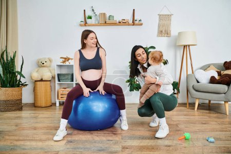 Una joven y hermosa madre se sienta graciosamente en una bola azul, sosteniendo tiernamente a su bebé mientras recibe orientación de su entrenador de crianza..