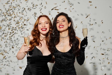 Two beautiful women in elegant attire clinking champagne flutes amid confetti.