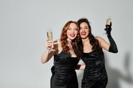 Two elegant women in black dresses joyfully toast with champagne flutes.