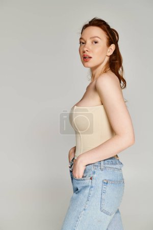 A woman in a tan top and blue jeans poses elegantly.
