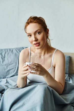 A woman in elegant attire sits on a bed, peacefully holding a cup of coffee.