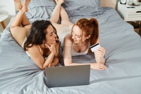 Foto de Dos mujeres en trajes elegantes, acostadas en una cama, felizmente enfocadas en un portátil. - Imagen libre de derechos